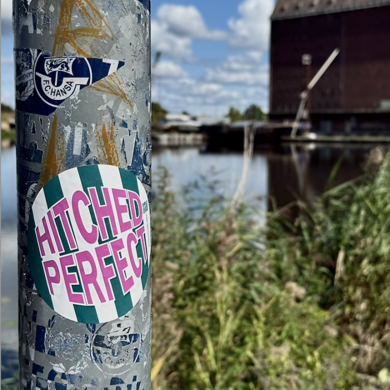Ein verzierter Pfosten mit einem Aufkleber „HITCHED PERFECT“ und eine Wasserlandschaft im Hintergrund.