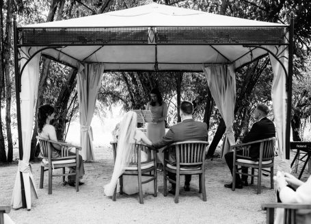 Vier Personen sitzen in einem Zelt unter Bäumen, im Gespräch mit Blick auf eine Wasserlandschaft.